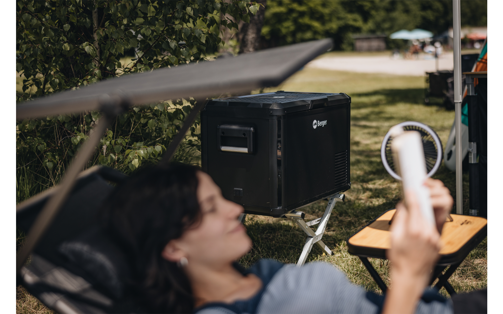 Person reclining in a lounge chair holding a smartphone; large black device on a stand, ring light and folding table in a park