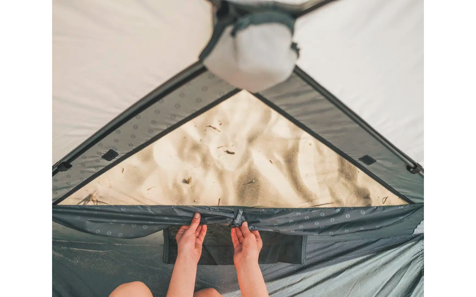 Blick von innen eines Zelts auf den Sandboden durch ein dreieckiges Moskitonetzfenster, zwei Hände öffnen die Fensterabdeckung