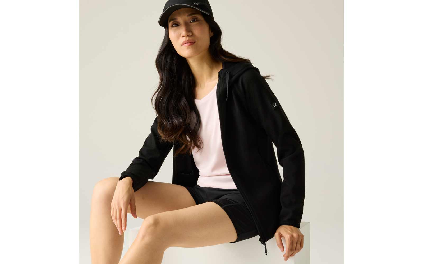 Person sitzt auf weißem Würfel, trägt schwarze Kapuzenjacke, schwarzes Cap, helles T‑Shirt und schwarze Shorts, lange dunkle Haare