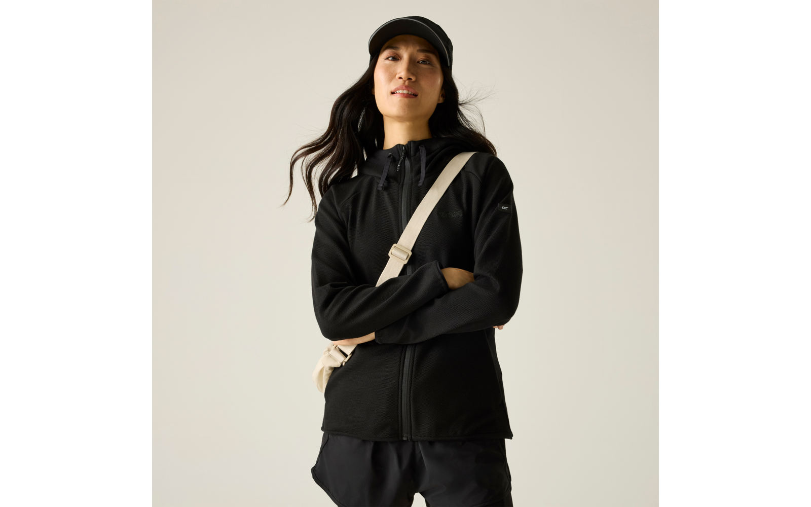 Person mit schwarzer Kappe, schwarzer Jacke mit Reißverschluss, sichtbarem beigem Umhängegurt, verschränkten Armen vor hellem Hintergrund