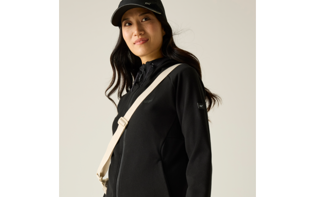 Person mit schwarzer Baseballkappe, schwarzer Jacke, beiger Umhängetasche und dunklen Haaren vor hellem Hintergrund