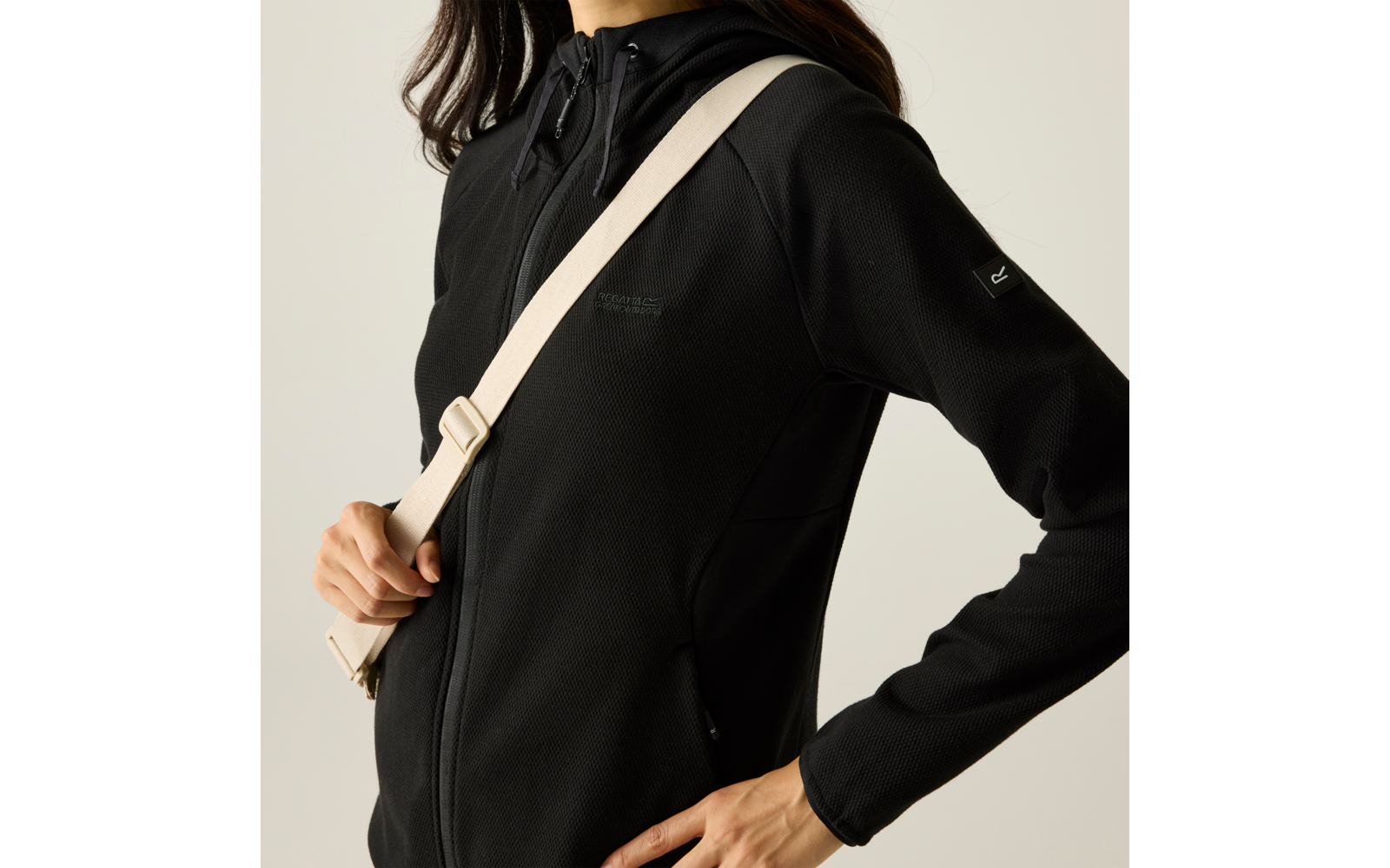 Teilweise sichtbarer Oberkörper einer Person in schwarzer Kapuzenjacke mit Reißverschluss, beigefarbener Schultergurt diagonal über der Brust, eine Hand hält den Gurt