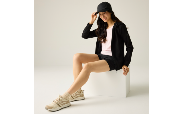 Frau mit schwarzer Kappe, schwarzer Jacke, rosafarbenem T-Shirt und schwarzen Shorts sitzt auf weißem Würfel, beige Sneakers