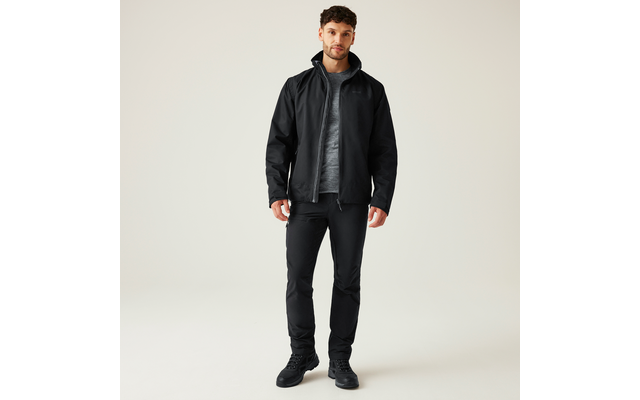 Ein Mann steht frontal vor hellem Hintergrund, trägt eine schwarze Jacke, ein graues T-Shirt, schwarze Hosen und schwarze Schuhe