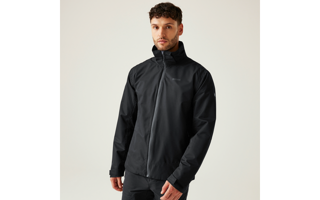 Ein Mann steht frontal vor hellem einfarbigem Hintergrund, trägt eine schwarze, hochgeschlossene Jacke mit Reißverschluss und schwarze Hose, Hände seitlich, blickt zur Kamera