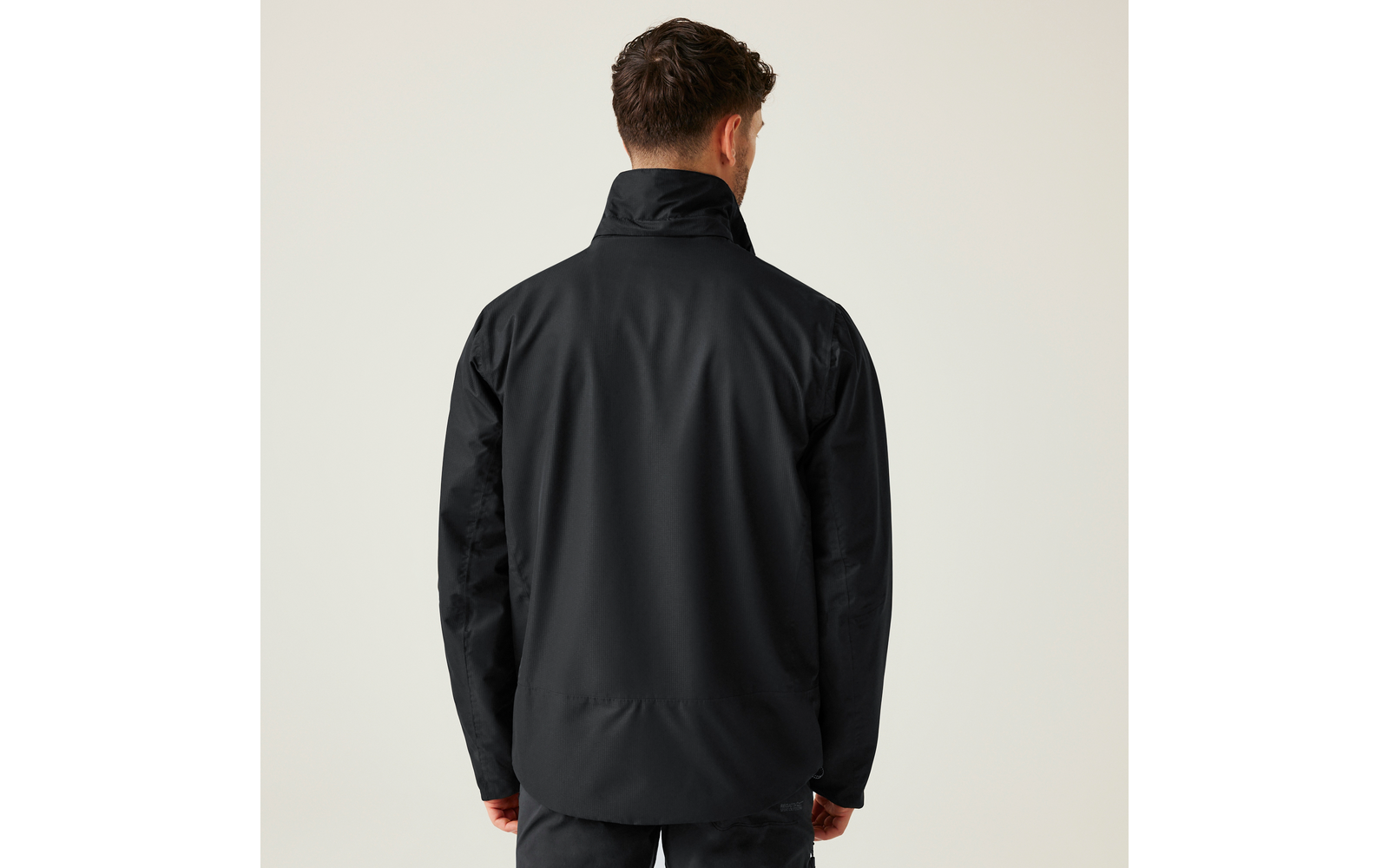 Person von hinten, trägt eine schwarze Jacke mit hohem Kragen und eine dunkle Hose vor hellem einfarbigem Hintergrund