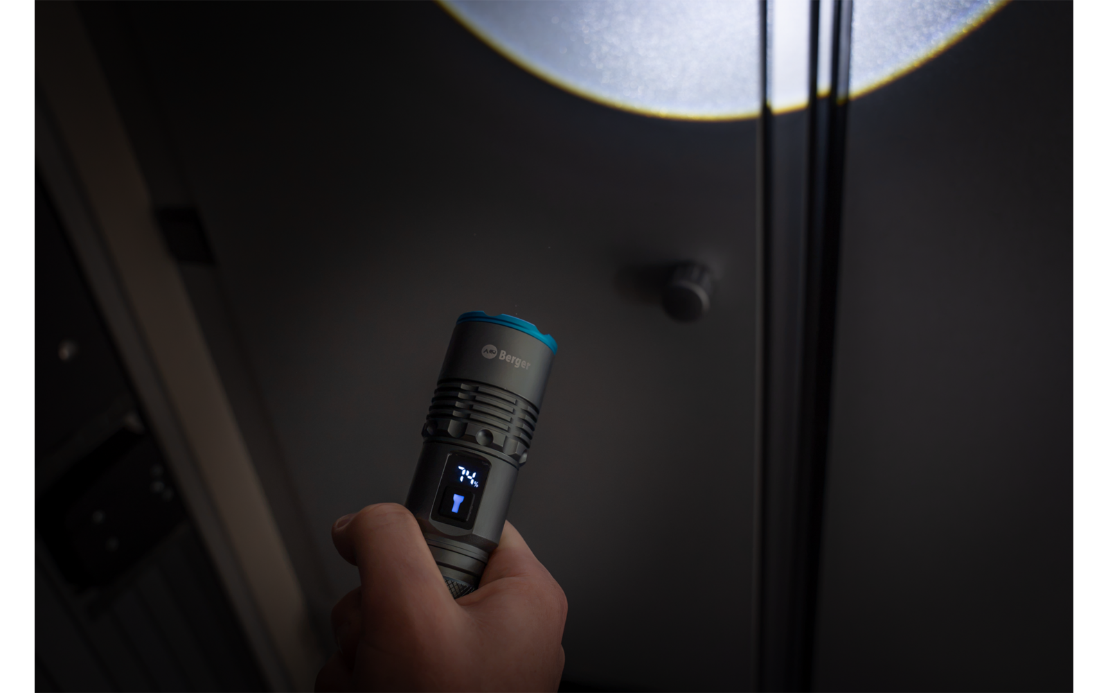 Hand hält eine schwarze Taschenlampe mit blauem Ring, der Lichtkegel bildet einen runden Fleck an einer grauen Decke neben einem kleinen runden Gerät