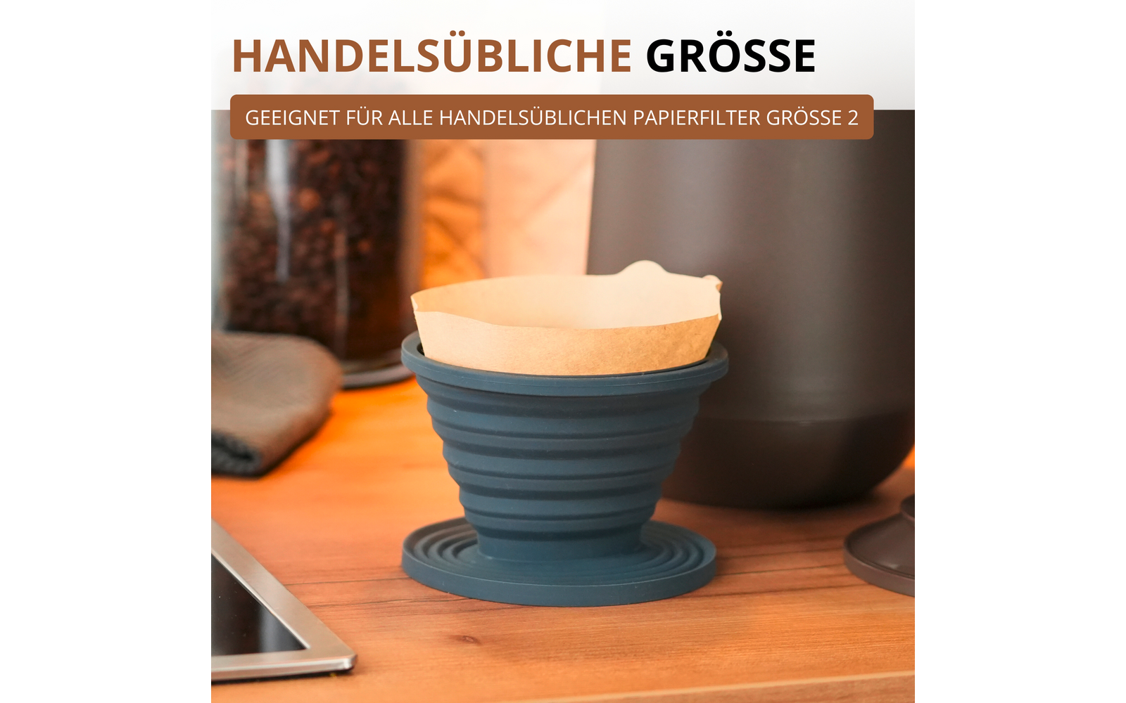 Support de filtre pliable en silicone bleu avec filtre en papier brun sur un plan de travail en bois ; en arrière-plan un bocal en verre de grains de café, une bouilloire foncée, un torchon plié et un coin d'ordinateur portable ; texte superposé « HANDELSÜBLICHE GRÖSSE » et « GEIGNET FÜR ALLE HANDELSÜBLICHEN PAPIERFILTER GRÖSSE 2 »