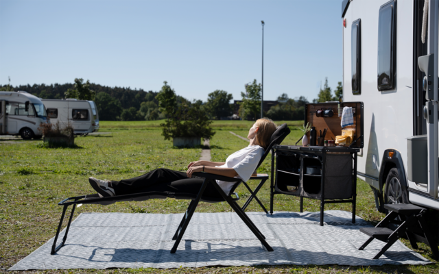 Person liegt auf Liegestuhl neben Wohnmobil auf Außenteppich, im Hintergrund weitere Wohnmobile und Wiese
