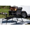 Schwarzer Liegestuhl mit Fußteil auf gemustertem Outdoor-Teppich, daneben weißer Wohnwagen mit offenem Fach und kleinem Klapptisch mit Geschirr, Gras im Hintergrund