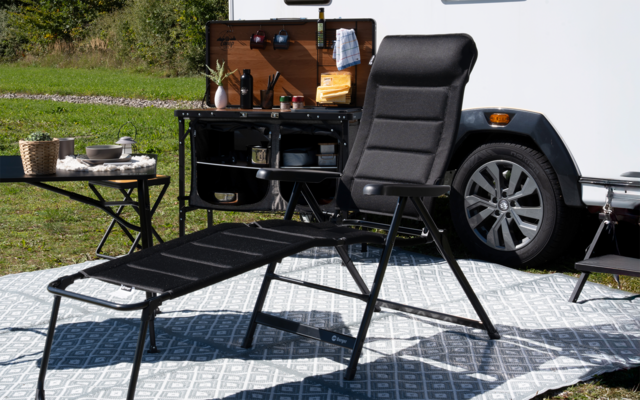 Schwarzer Liegestuhl mit Fußteil auf gemustertem Outdoor-Teppich, daneben weißer Wohnwagen mit offenem Fach und kleinem Klapptisch mit Geschirr, Gras im Hintergrund