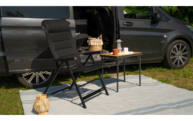 Schwarzer Klappstuhl und Campingtisch mit Kaffeetasse und Kerze vor offenem grauem Auto auf einer hellen Decke im Freien