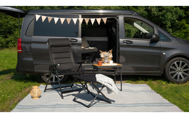Aufgeklappter grauer Campingbus mit geöffneten Türen, davor zwei Campingstühle und ein kleiner Tisch auf einer Decke im Gras