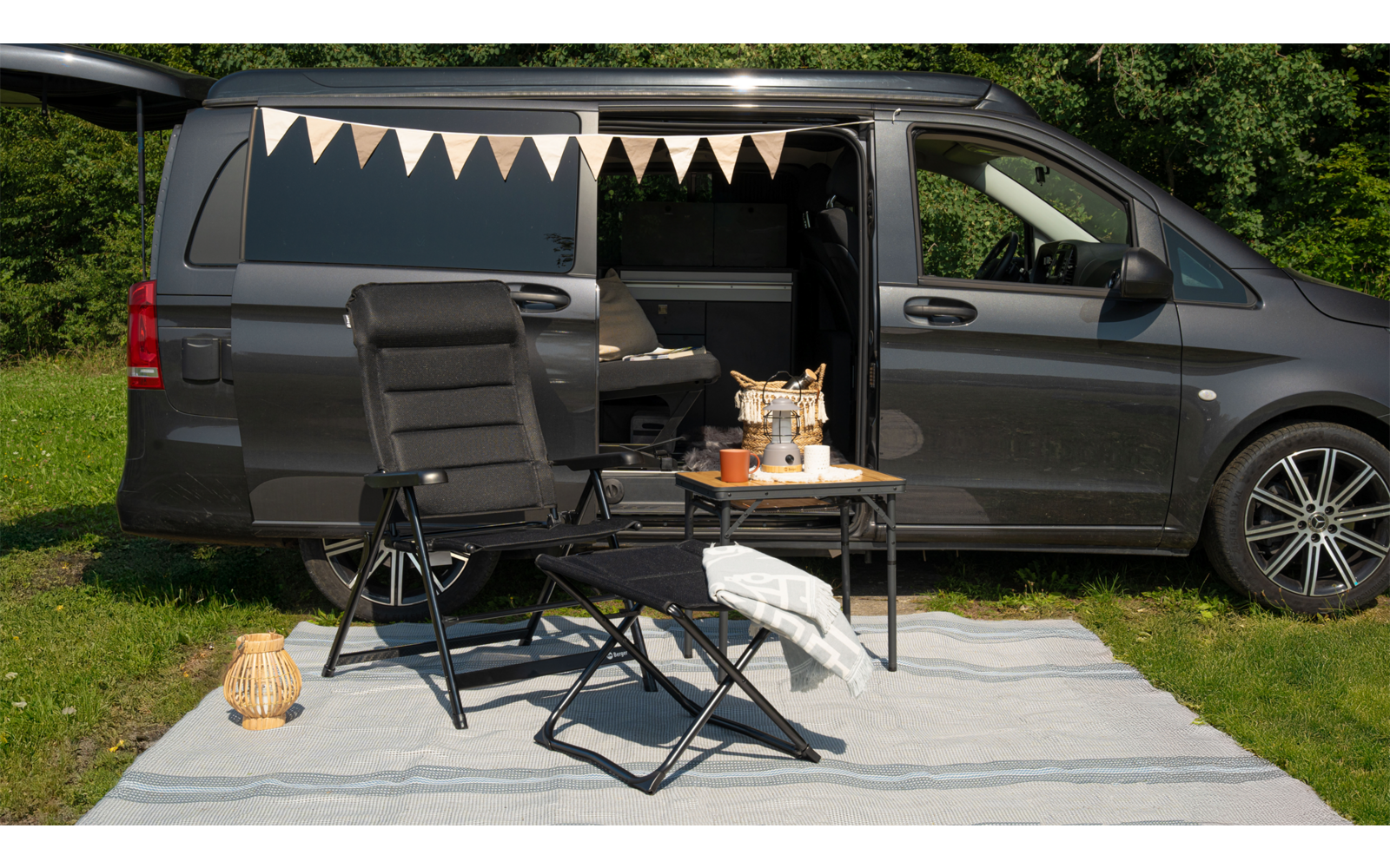 Aufgeklappter grauer Campingbus mit geöffneten Türen, davor zwei Campingstühle und ein kleiner Tisch auf einer Decke im Gras