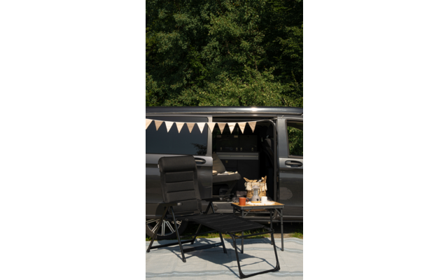 Zwei schwarze Klappstühle und ein kleiner Tisch mit Campinggeschirr vor einem geöffneten schwarzen Van, im Hintergrund Bäume