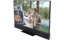 Mann und Frau stehen nebeneinander vor einem Wohnmobil in einem bewaldeten Gebiet, halten jeweils ein Stück Wassermelone; geöffnete Wohnmobil-Seite mit Campingausrüstung, Hängematte zwischen Bäumen