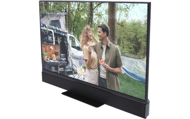 Mann und Frau stehen nebeneinander vor einem Wohnmobil in einem bewaldeten Gebiet, halten jeweils ein Stück Wassermelone; geöffnete Wohnmobil-Seite mit Campingausrüstung, Hängematte zwischen Bäumen