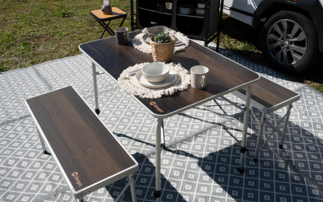 Faltbarer Tisch mit zwei Bänken auf einer grauen, geometrisch gemusterten Matte; auf dem Tisch Teller, Schale, Tasse, geflochtener Korb mit Pflanze und ein kleines Tuch; im Hintergrund Seitenwand und Reifen eines Fahrzeugs