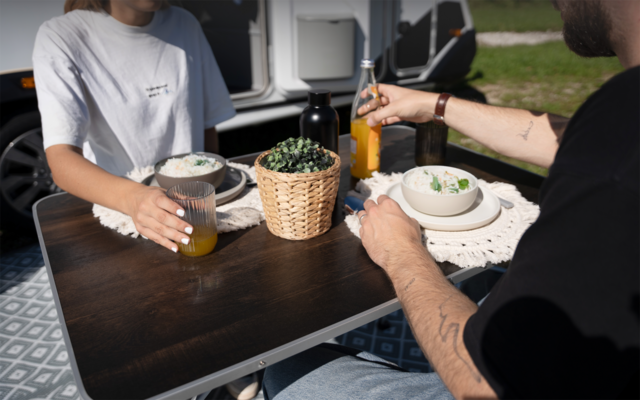 Zwei Personen an einem kleinen dunkelbraunen Tisch vor einem Wohnwagen; auf dem Tisch zwei Gläser Saft, eine Saftflasche, zwei Schalen mit Essen, ein Rattankorb mit Pflanze und weiße Platzsets