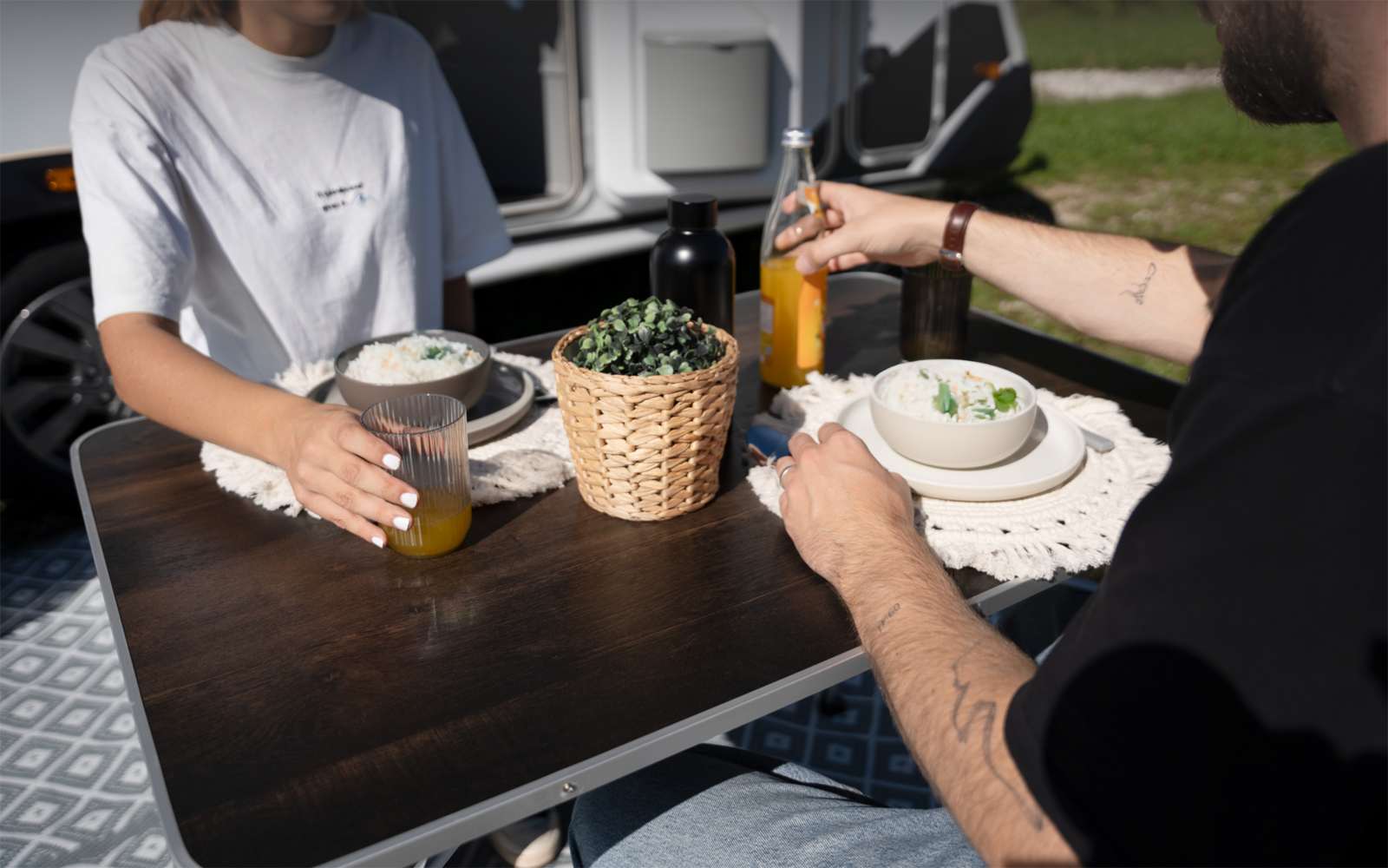 Zwei Personen an einem kleinen dunkelbraunen Tisch vor einem Wohnwagen; auf dem Tisch zwei Gläser Saft, eine Saftflasche, zwei Schalen mit Essen, ein Rattankorb mit Pflanze und weiße Platzsets