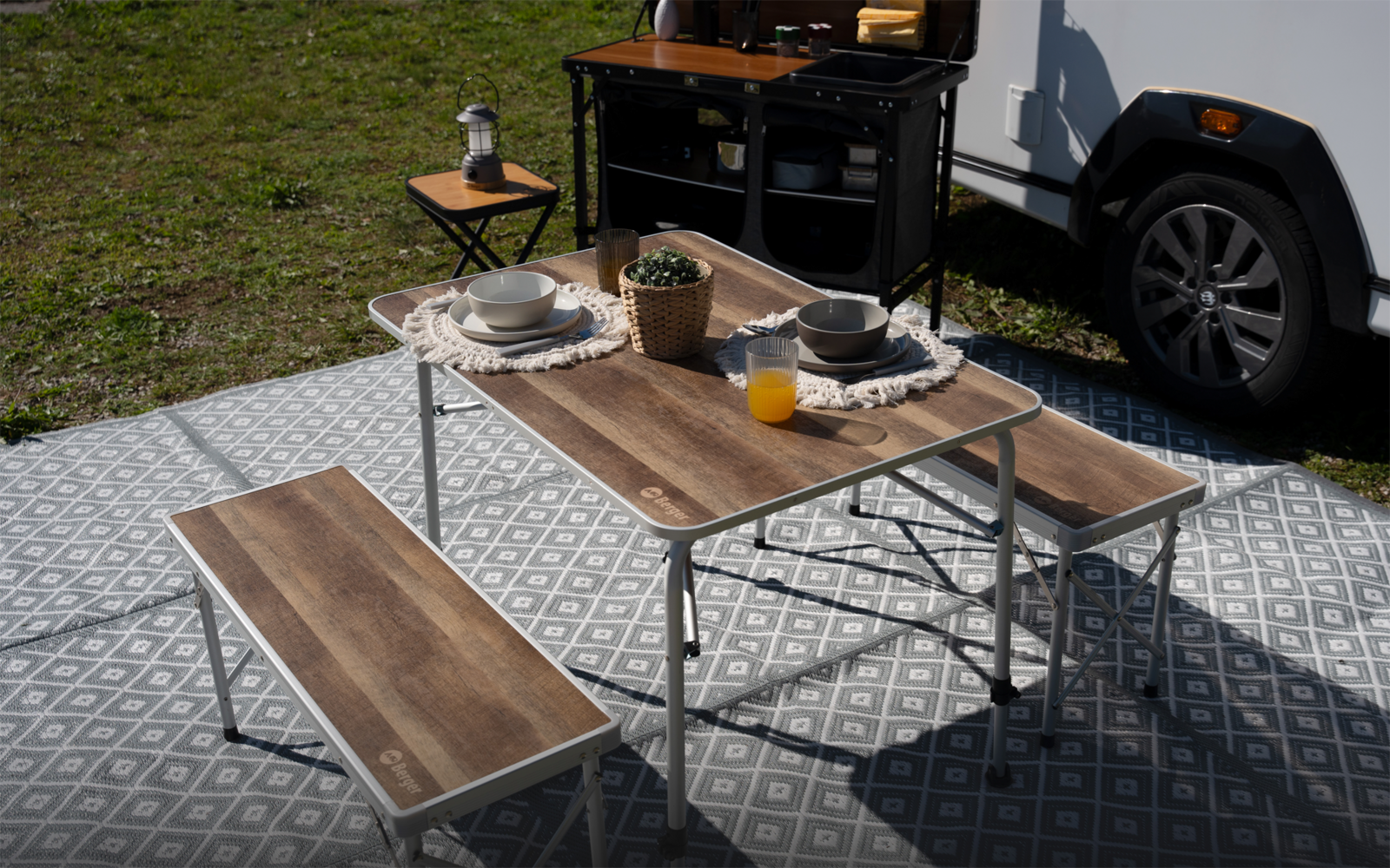 Klappbarer Holztisch mit zwei Bänken auf grauem Musterteppich; auf dem Tisch zwei Teller, Schüsseln, Tassen, ein Glas Orangensaft und eine kleine Topfpflanze; im Hintergrund Campingküche, Wohnwagen und Rasen