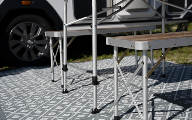 Faltbarer Tisch und Hocker mit Metallbeinen auf gemusterter Außenmatte, im Hintergrund ein Fahrzeugreifen