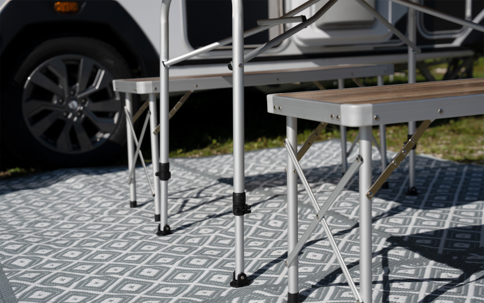 Faltbarer Tisch und Hocker mit Metallbeinen auf gemusterter Außenmatte, im Hintergrund ein Fahrzeugreifen