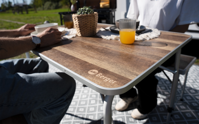Holztisch mit 'Berger'-Logo, Glas Orangensaft, weiße Tasse, geflochtener Topf mit Pflanze, zwei Personen (nur Arme und Beine sichtbar), gemusterte Matte, Gras im Hintergrund