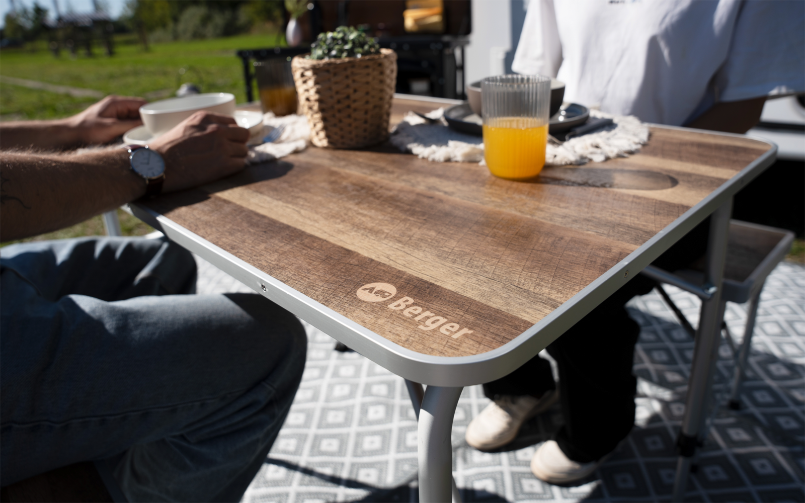 Holztisch mit 'Berger'-Logo, Glas Orangensaft, weiße Tasse, geflochtener Topf mit Pflanze, zwei Personen (nur Arme und Beine sichtbar), gemusterte Matte, Gras im Hintergrund