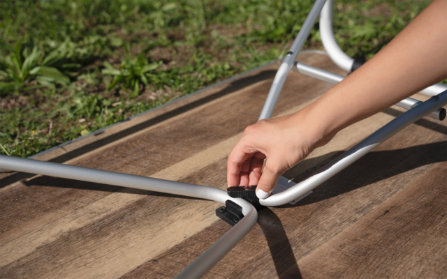 Hand berührt silberne Metallstangen mit schwarzem Verbinder auf Holzplanken, Gras im Hintergrund