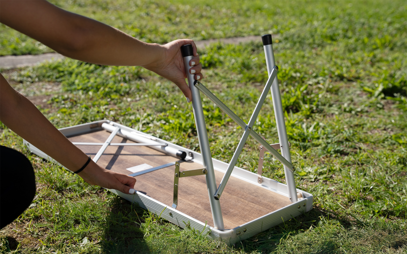 Two hands unfolding metal legs of a folding table with wooden top and white frame on grass