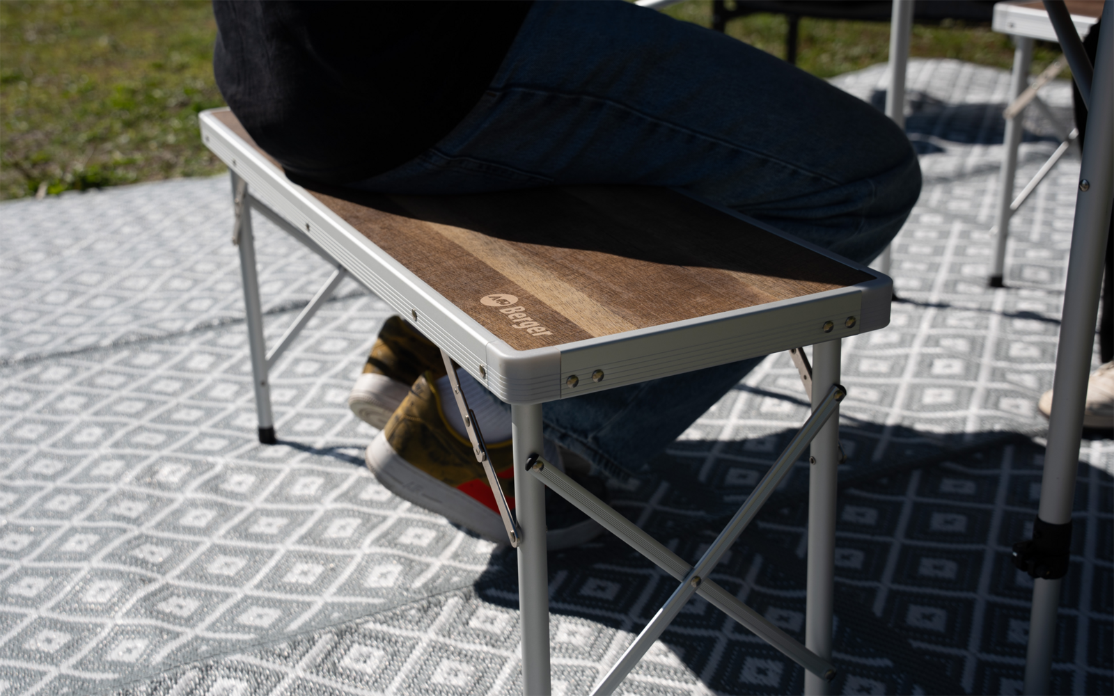 Person seated on a folding bench with wooden top and aluminum frame on a patterned outdoor mat; visible legs in blue jeans and brown shoes