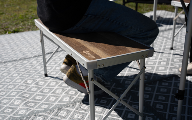 Person seated on a folding bench with wooden top and aluminum frame on a patterned outdoor mat; visible legs in blue jeans and brown shoes