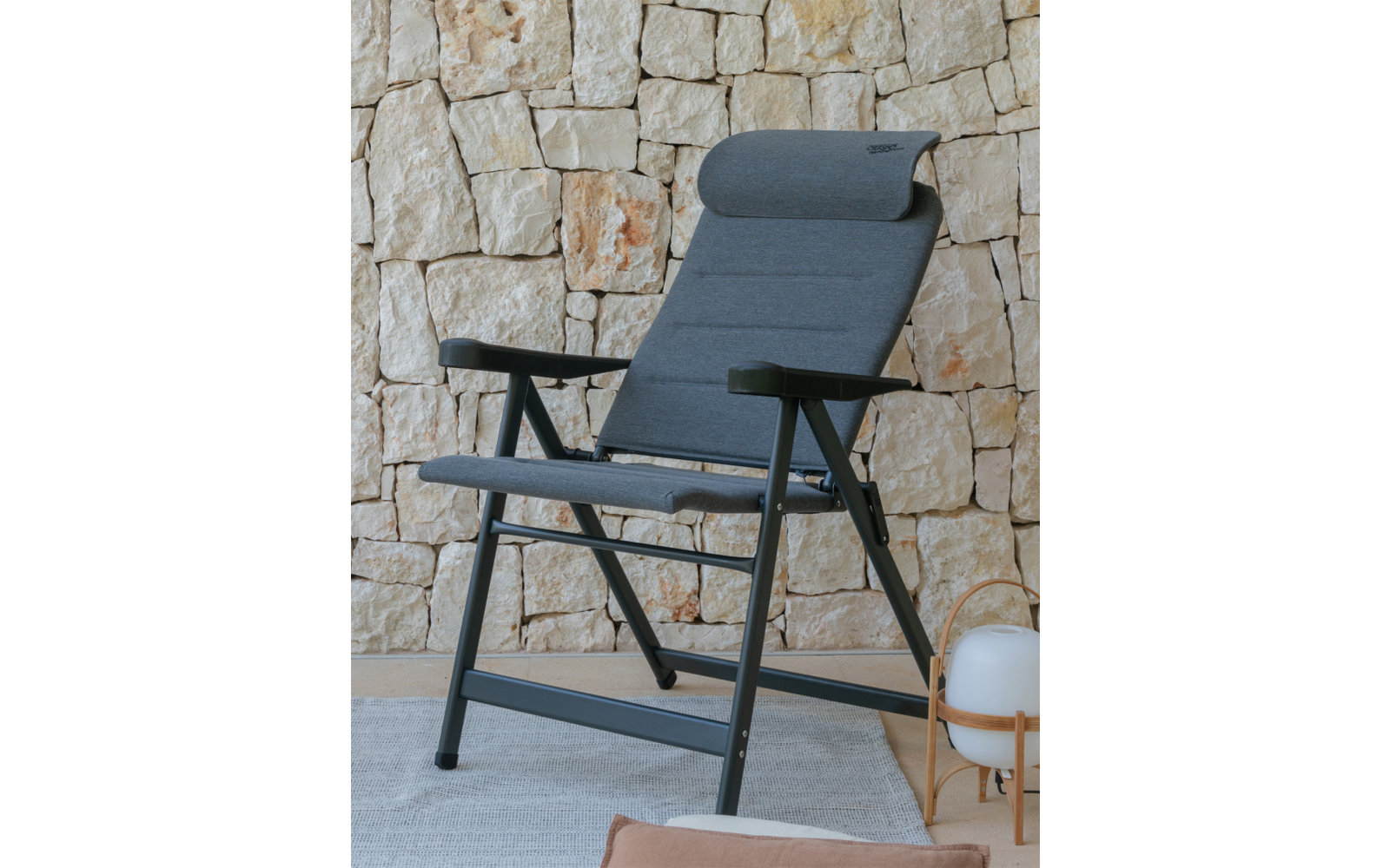 Grauer gepolsterter Liegestuhl mit Nackenrolle und schwarzem Metallgestell vor einer hellen Natursteinwand, heller Teppichboden, rechts eine runde weiße Lampe auf Holzgestell, vorne ein beiges Kissen