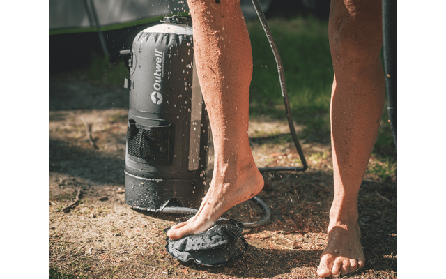 Untere Beine und nackte Füße einer Person neben einem zylindrischen Duschbehälter mit aufspritzendem Wasser auf trockenem Gras; ein Duschschlauch sichtbar