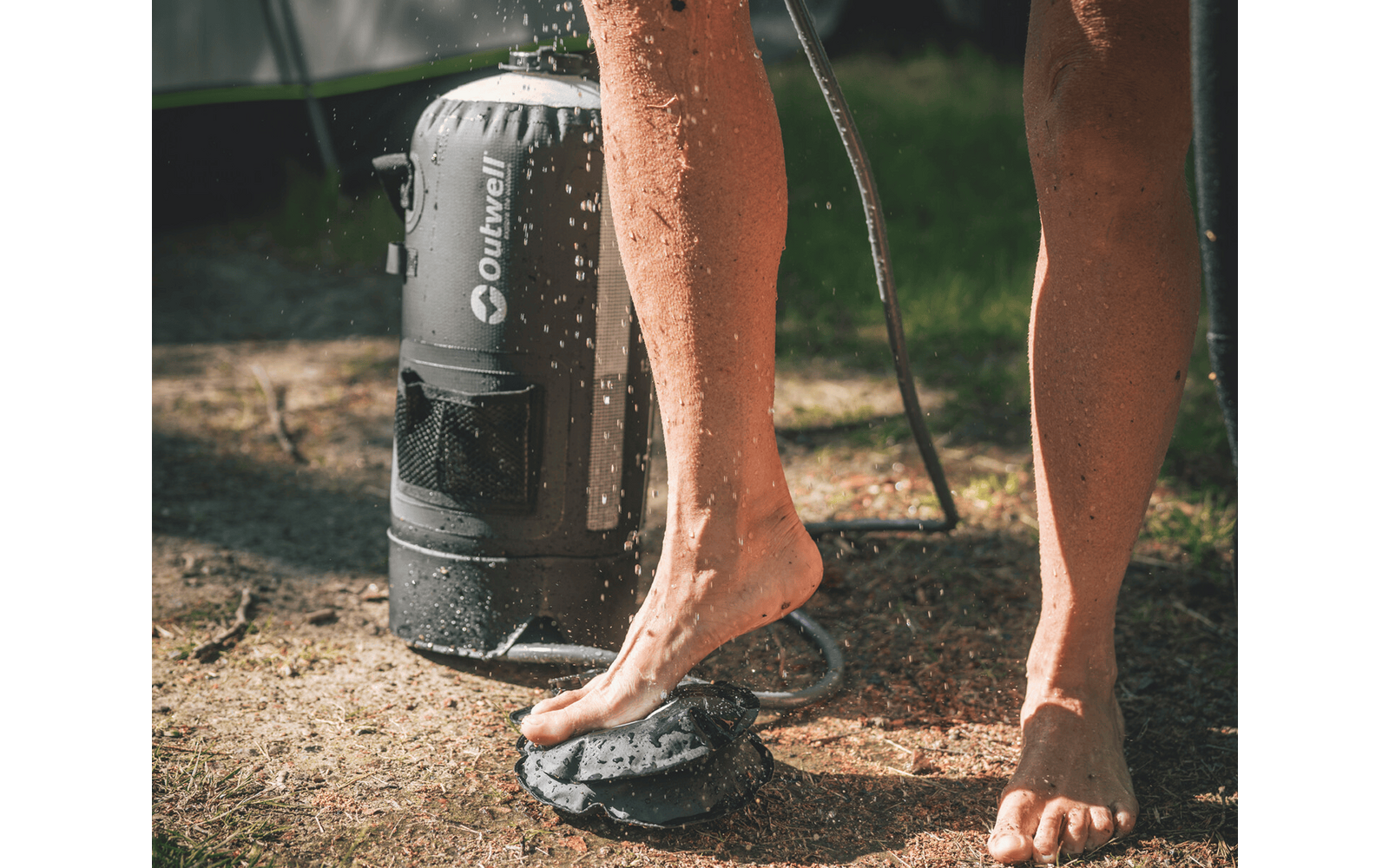 Untere Beine und nackte Füße einer Person neben einem zylindrischen Duschbehälter mit aufspritzendem Wasser auf trockenem Gras; ein Duschschlauch sichtbar