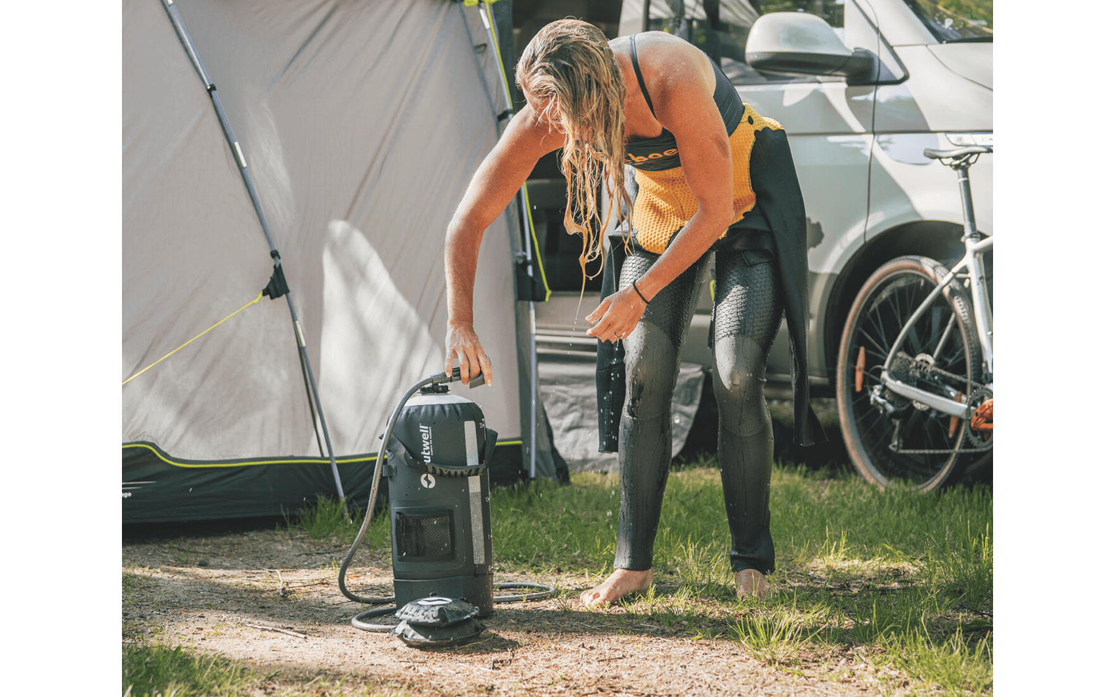 Person mit nassen, schulterlangen blonden Haaren, trägt ein gelbes Handtuch um den Oberkörper und schwarze Neoprenhose, steht barfuß auf Gras und berührt eine tragbare Außendusche; im Hintergrund Zelt, weißes Fahrzeug und Fahrrad