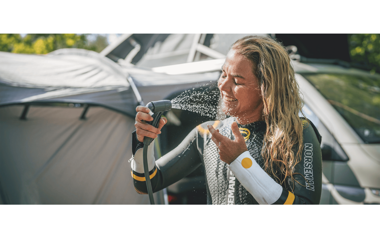 Person mit langen blonden Haaren in Neoprenanzug spritzt sich mit Gartenschlauch Wasser ins Gesicht, Zelt und Fahrzeug im Hintergrund