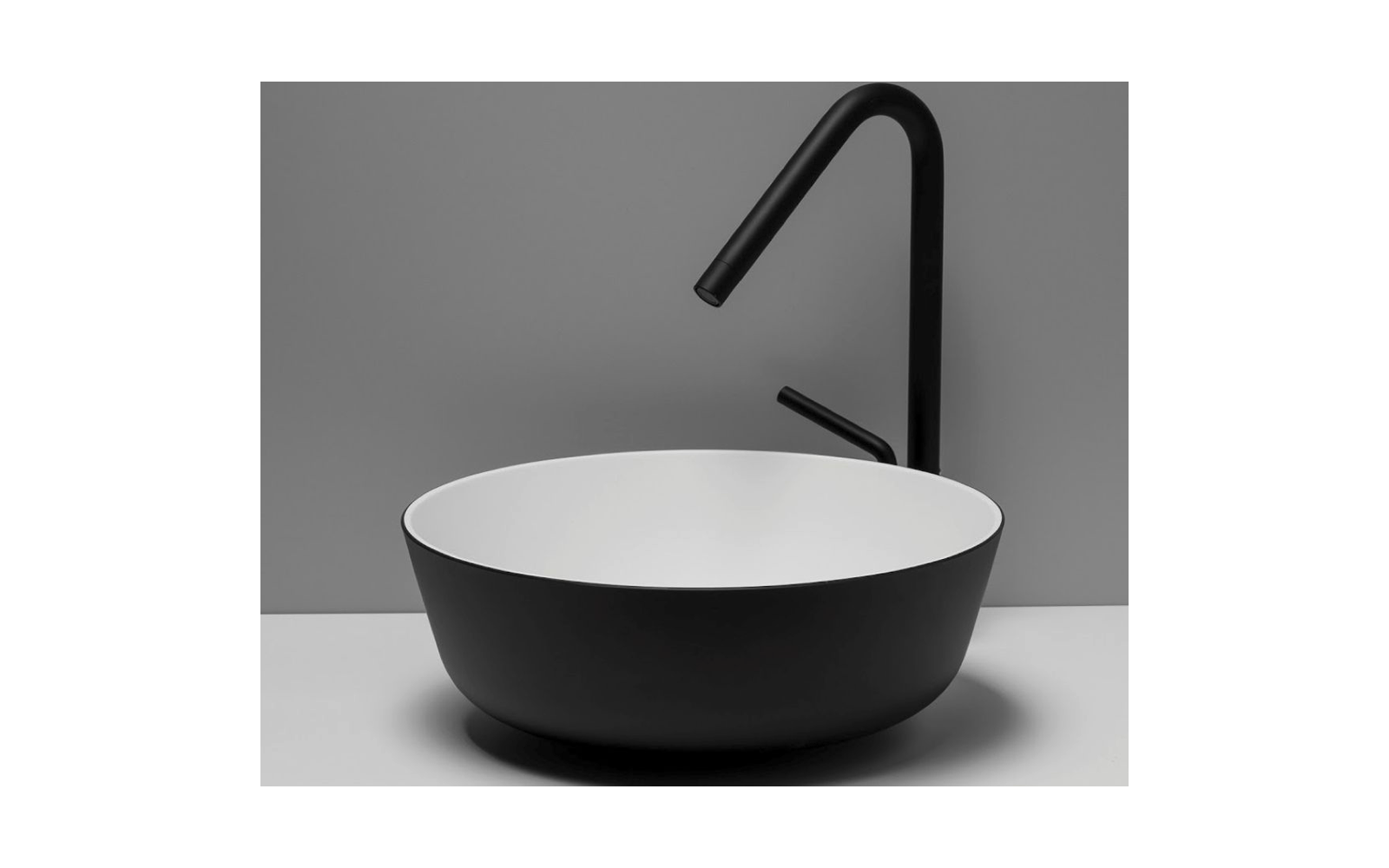 Lavabo rotondo con esterno nero e interno bianco su un piano chiaro; alto rubinetto nero con beccuccio curvo e leva; parete grigia sullo sfondo