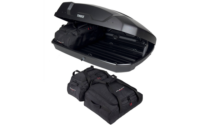 Geöffnete schwarze Dachbox auf weißem Hintergrund, im Inneren eine schwarze Reisetasche, drei weitere schwarze Reisetaschen liegen davor