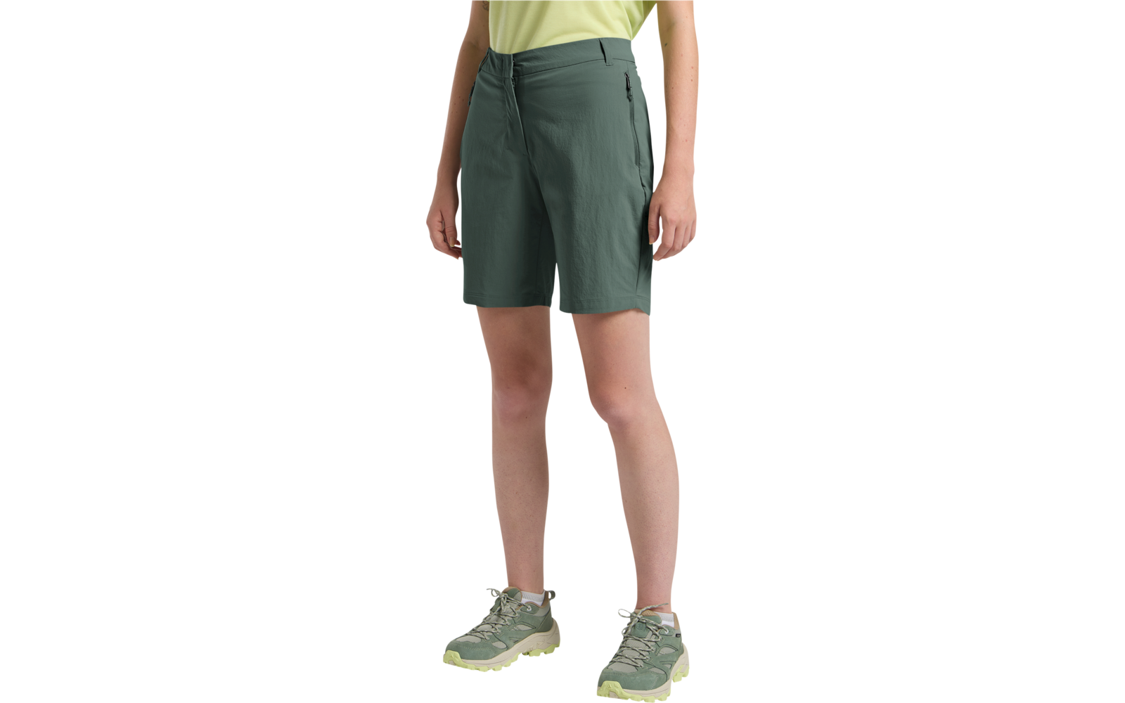 Untere Körperhälfte einer stehenden Person mit dunkelgrünen knielangen Shorts, gelbem Oberteil sichtbar am Bund, hellgrünen Sportschuhen mit hellgelben Sohlen und seitlich hängenden Händen