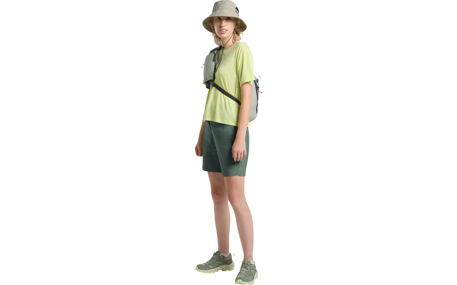 Personne debout, légèrement tournée, portant un chapeau seau beige, un t-shirt vert clair à manches courtes, un short vert foncé, un petit sac bandoulière vert et des baskets vert clair