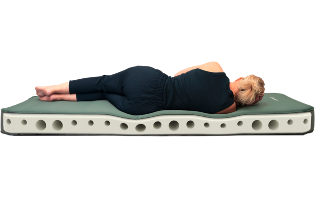 Person with short blond hair wearing a black top and black leggings lies on their side on a green pad on a white foam mattress with round holes