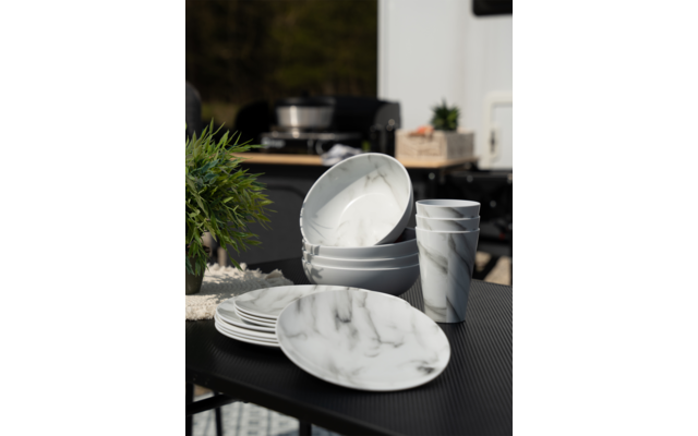 Mehrere weiße Geschirrteile mit grauem Marmor-Muster auf dunklem Tisch, im Hintergrund unscharfe grüne Pflanze