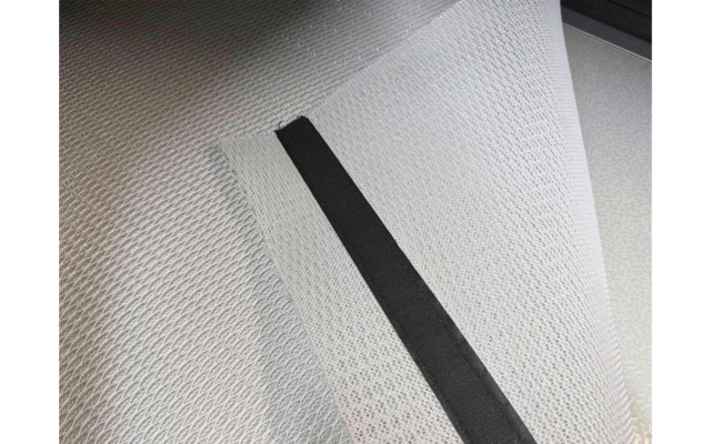 Lichtgrijze gestructureerde mat met een opgetilde hoek, zwarte strook langs de gevouwen rand, ronde metalen bevestiging op de vloer rechts