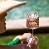 Person mit Sonnenhut hält ein facettiertes Weinglas mit roséfarbenem Getränk; im Hintergrund unscharfer Pool und Rasen