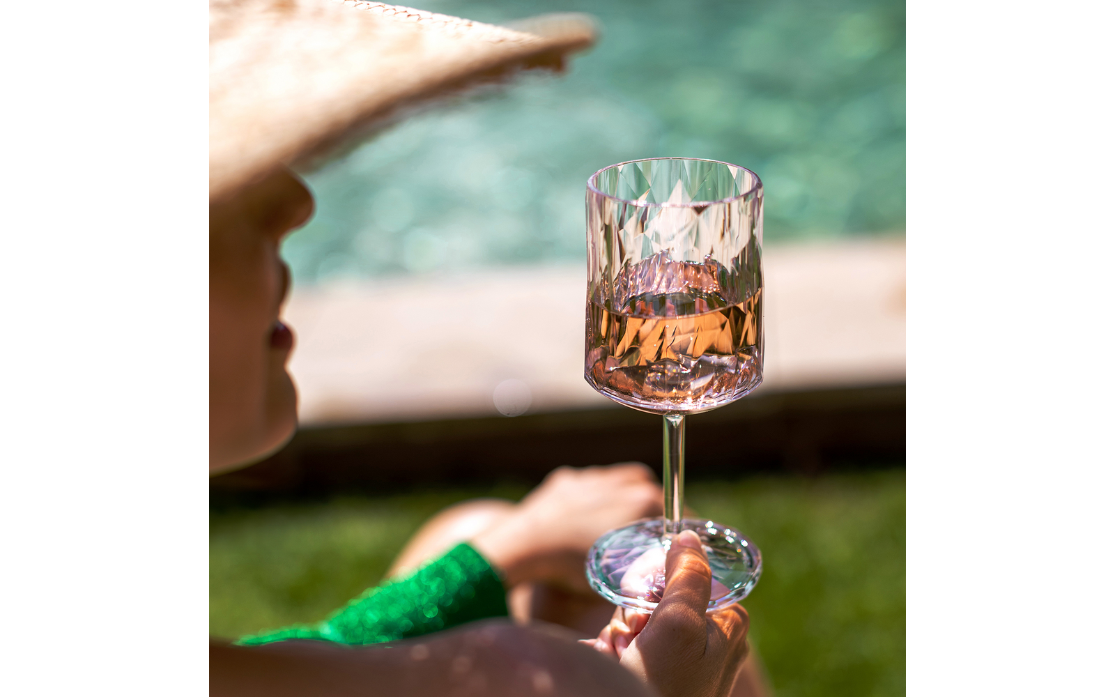 Person mit Sonnenhut hält ein facettiertes Weinglas mit roséfarbenem Getränk; im Hintergrund unscharfer Pool und Rasen