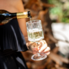 Hand hält ein Weinglas, in das eine Flasche Schaumwein eingeschenkt wird; unscharfe Holzscheite im Hintergrund