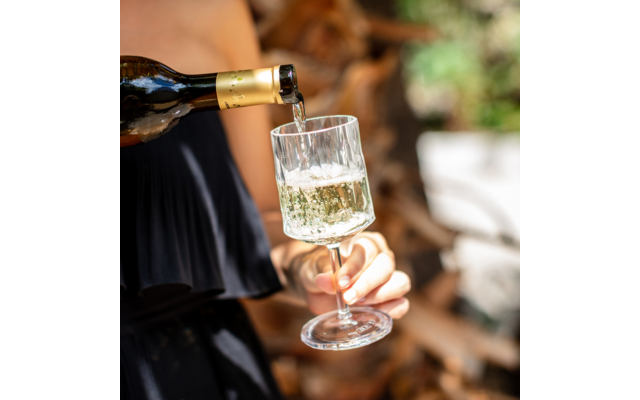 Hand hält ein Weinglas, in das eine Flasche Schaumwein eingeschenkt wird; unscharfe Holzscheite im Hintergrund