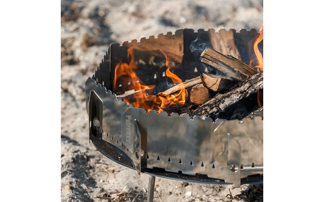 Metallfeuerschale auf kurzen Beinen mit brennenden Holzscheiten und Flammen auf sandigem Untergrund